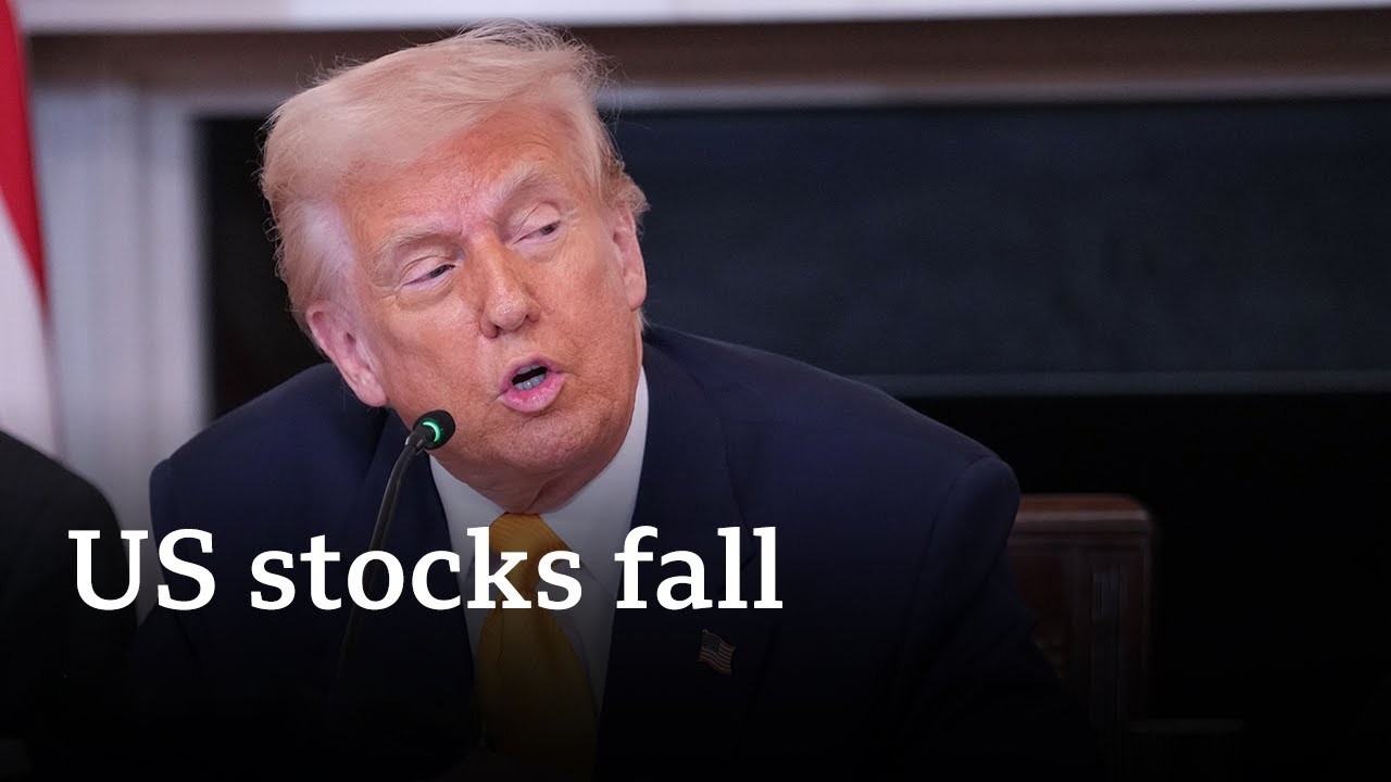 A man in a dark suit and yellow tie speaking into a microphone, with the headline "US stocks fall" overlaid on the image.